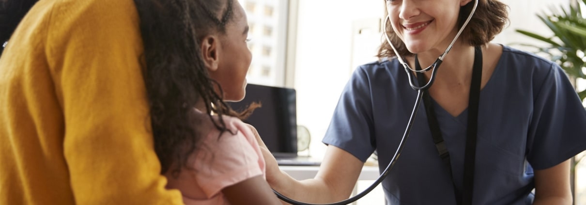 Female Pediatrician Wearing Scrubs Listening To Girls Chest With Stethoscope In Hospital Office
